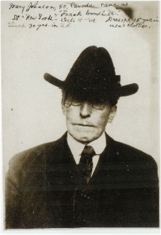In this photo, Frank wears a black suit and tie, with a white collar. Frank is light-skinned and has a dimpled chin and a white moustache, and wears circular spectacles with a small chain leading from the right lens back toward the ear. The black top hat on Frank's head is made of a heavy material with a wide brim and sits low on Frank's forehead so that the forehead is fully obscured and Frank's eyes are shadowed. Sherman's hand-written caption, written on the photo above Frank's head, says "Mary Johnson, 50, Canada came as 'Frank Woodhull' SS New York Oct. 4 '08. Lived 30 yrs. in U.S. Dressed 15 yrs. in men's clothes."