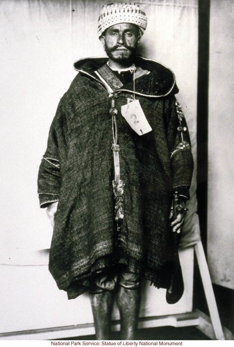 This image is labeled "North African Immigrant," and the picture shows a seemingly middle-aged, dark-skinned person with a beard, a knit or woven hat, and a large hooded jacket, frayed at the bottom and closed with buttons at the front. The subject's legs are bare. The subject also has a tag affixed to his jacket with the number "2" printed on it.