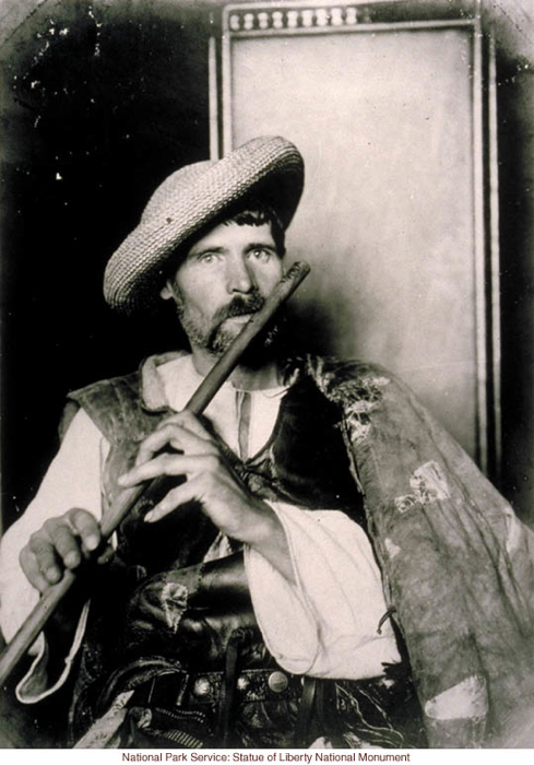 This image is labeled "Eastern European Immigrant," and the picture shows a young person with a beard, a woven hat with a wide brim angled to the side of the head. The subject wears a sort of cape, a vest, and a white shirt tied at the neck. The subject plays a flute that is held out in front of the subject. The subject seems quite tanned but not particularly dark-skinned.