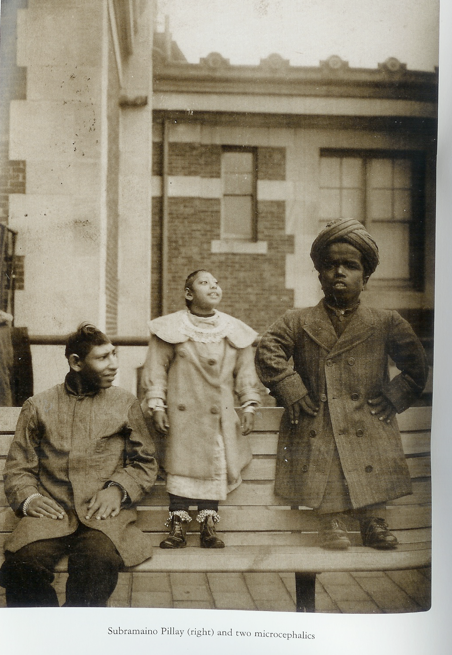 This image pictures three people on the roof of a building (a common backdrop for Sherman's photos). On the left, seated on a bench, there is a dark-skinned person with their head turned to our right, wearing a knee-length jacket. Beside him another dark-skinned person stands on the bench, so as to draw attention to the subject's comparatively smaller stature-the subject stands at about the same head-level as the seated person. This middle subject is wearing an ankle-length jacket with fur trim, and their chin is raised in the air. On the far right, there is a third subject, also dark-skinned and standing on the bench and just slightly taller than the person in the middle of the picture. This final subject wears a turban, and their head is larger than those of the other two subjects. This final subject also wears an ankle-length coat and holds their hands on their waist. "The image is labeled Subramaino Pillay (Right) and Two Microcephalics."