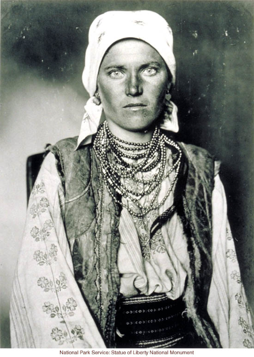 This image shows a young person (the title genders the subject as female) wearing a white headscarf, a vest, a blouse with floral stitching down the sleeves and front, and many beaded necklaces. The subject gazes directly at the camera. Though the photo is black and white, the eyes have an unusual, light color that has a striking effect. The subject's skin is tanned. 