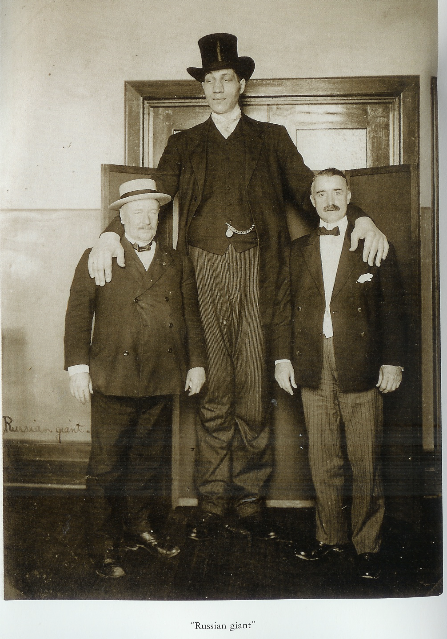 The "Russian Giant" stands with hands on the shoulders of two white people in suits, whose heads reach only to the Russian immigrant's waist. The subjects in suits each have moustaches. The Russian wears a top hat and tuxedo with tails. The chain of a pocket watch is visible where the suit jacket is open. There is a set of double doors behind the subject, and the Russian is clearly taller than this doorway.
