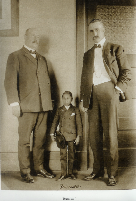 The "Burmese" man stands between two white subjects in suits, the same two subjects from the "Russian Giant" picture. The Burmese subject has dark skin and stands at waist-height of the subjects on either side, and holds a top hat. The Burmese subject wears a double-breasted suit.
