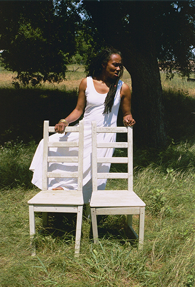 A woman in a white sundress is standing in front of a tree and looking off to the side. There are two wooden chairs in front of her, and she’s resting one hand on the top of each chair.