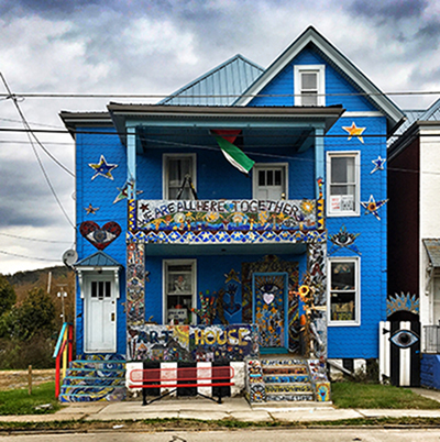 Image of a two-story home painted bright blue and covered in art from top to bottom. The words “WE ARE ALL HERE TOGETHER” adorn the mosaic artwork on the second-floor balcony.