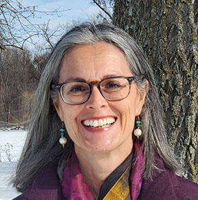 A close-up headshot of the author smiling against a tree trunk with a snowy background, in a brightly colored winter jacket, wearing dark-rimmed glasses and dangling earrings