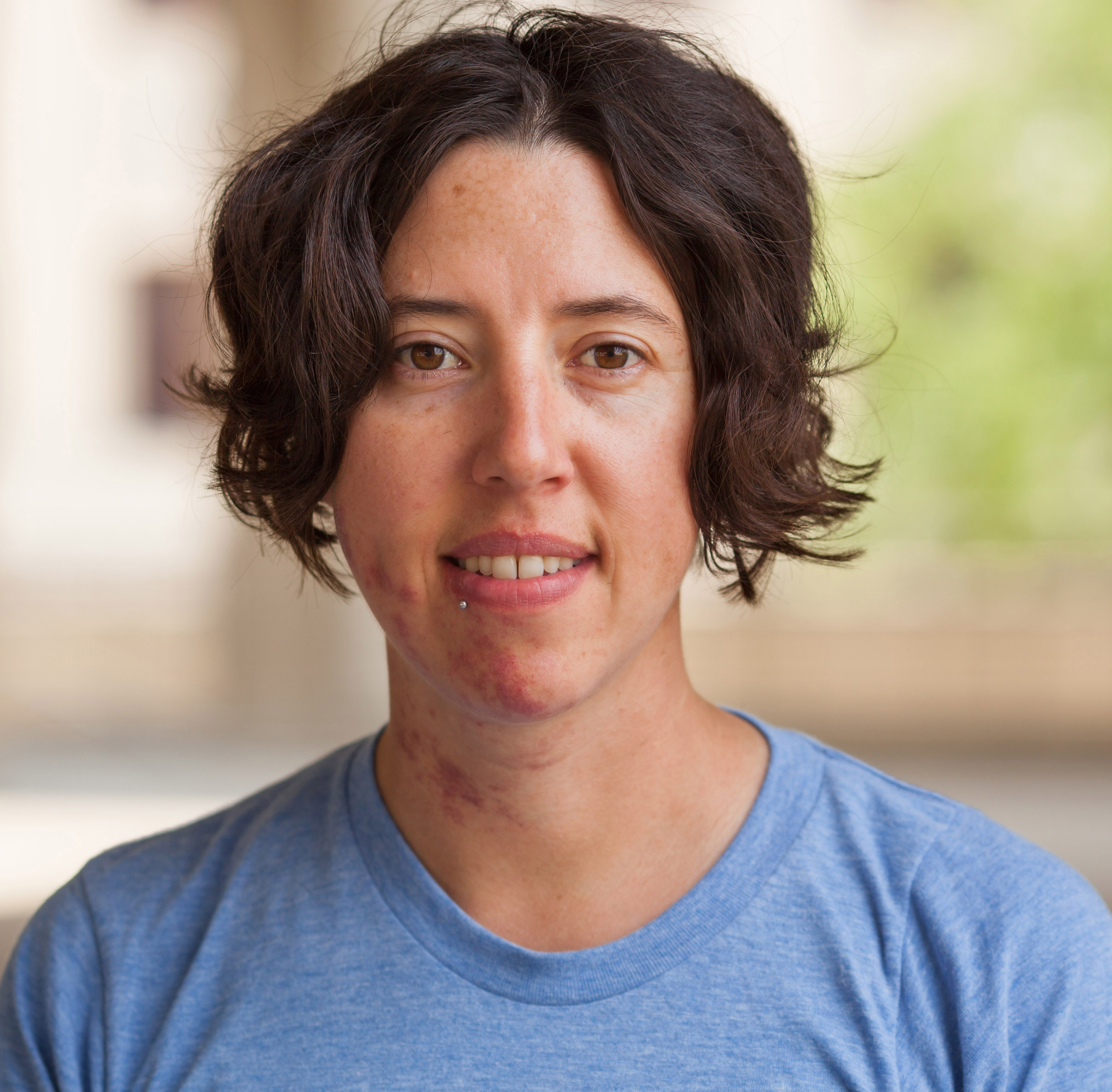 headshot of Karma Chavez, depicting her looking directly at the camera with a slight smile and open expression, with short, wavy dark brown hair, and wearing a blue t-shirt